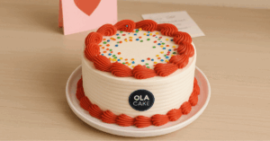 Pastel decorado con crema blanca y bordes rojos, adornado con confites de colores, sobre una mesa de madera clara. Al fondo, una tarjeta rosada con un corazón rojo simboliza amor y celebración, ideal para cumpleaños o aniversarios en Cuba.